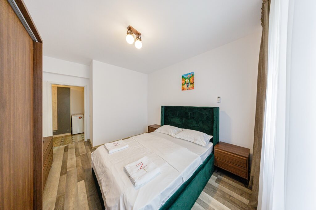 Dormitor de apartament cu pat matrimonial, tăblie verde și dulap, cu acces la baie.