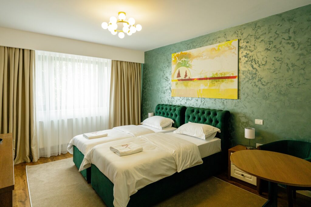 Cameră twin cu două paturi alăturate, tăblie capitonată verde, candelabru auriu și tablou decorativ.
