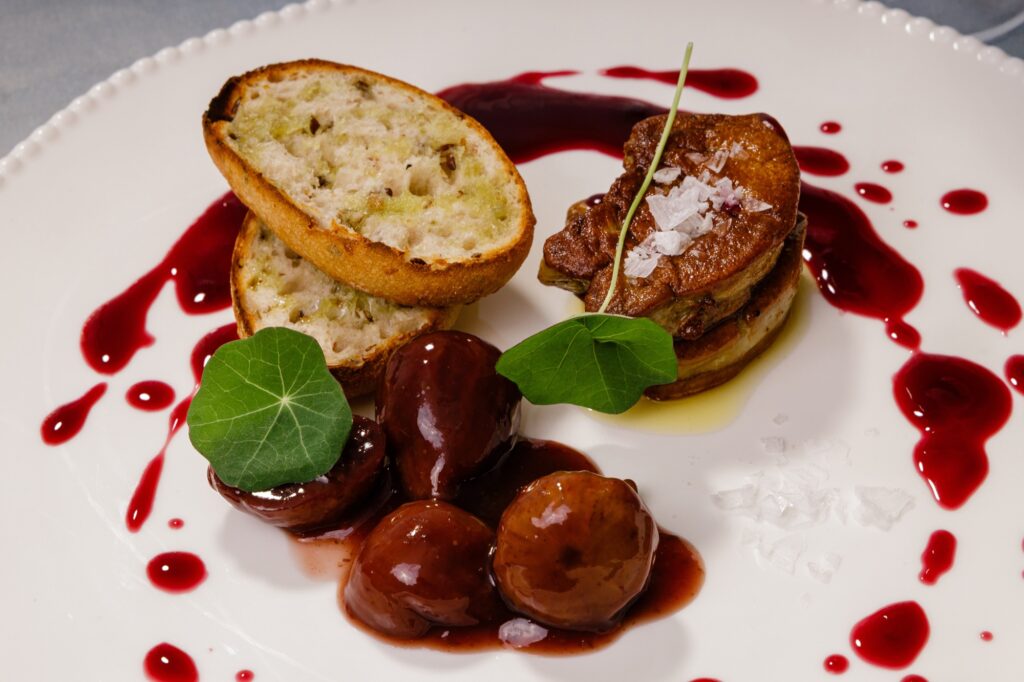Foie gras la tigaie cu crostini, vișine în sos de vin roșu și flori comestibile, pe farfurie albă