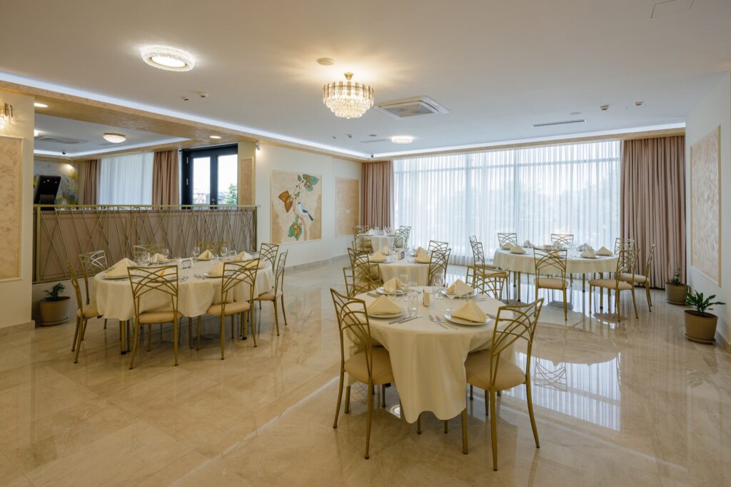 Salon restaurant cu mese rotunde aranjate elegant, scaune aurii și tablou decorativ pe perete.