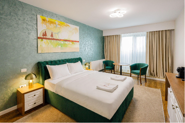 Camera matrimoniala din hotelul IN Grand Hotel&SPA, amenajata cu pat, tablou, noptiere, fotolii,masa si nuante de verde si auriu